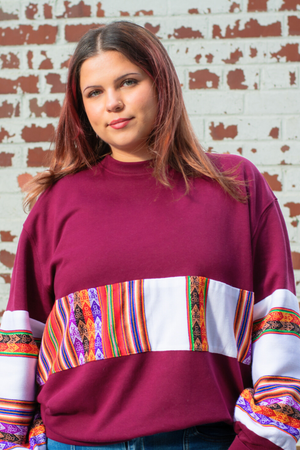 Woman wearing a Latin-inspired, ethically-made, burgundy sweater with colorful patterned sleeves made with Aguayo fabric from Peru against a brick wall.