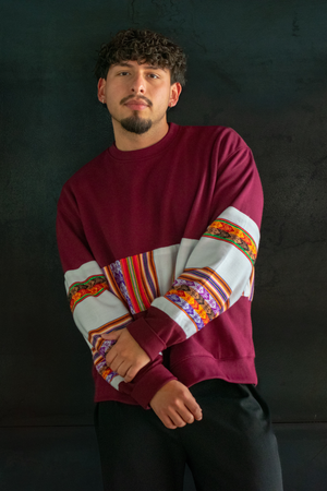 Man wearing a Latin-inspired, ethically-made, burgundy sweater with colorful patterned sleeves made with Aguayo fabric from Peru against a dark background.