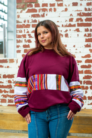 Woman wearing a Latin-inspired, ethically-made, burgundy sweater with colorful patterned sleeves made with Aguayo fabric from Peru against a brick wall.
