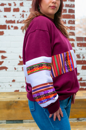 Close up of a woman wearing a Latin-inspired, ethically-made, oversized, burgundy sweater with colorful patterned sleeves made with Aguayo fabric from Peru against a brick wall