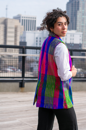 Side profile of a model wearing a Latin-inspired, artisan-made, semi-fitted, colorful vest handwoven in El Salvador with a collar over a white button-up shirt. 