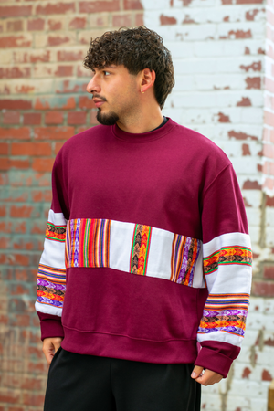 Man wearing a Latin-inspired, ethically-made, burgundy sweater with colorful patterned sleeves made with Aguayo fabric from Peru against a brick wall.